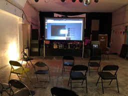 An events space with a stage with drum kit, a projector screen, chairs and no people.