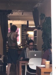 Coffee shop, a man works on an Apple Laptop, another man in a string vest looks at a menu on a blackboard.