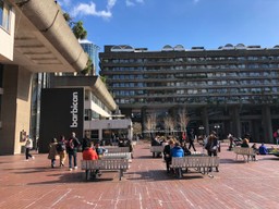 The Barbican Centre entrance. A large terrace with people picknicking, a two storey sign reads 'Barbican' in the background we can see what could best be described as the Ancient Hanging Gardens Of Babylon