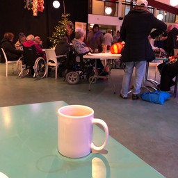 Old Ppeople in a cafe, some in wheelchairs, a Christmas tree is in the baackground, a cup of tea sits on a table in the foreground