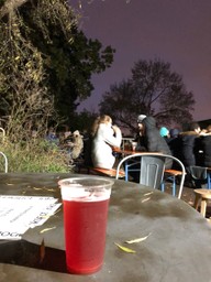Red/purple looking beer on a table, in the backround people drinking at a table outdoors, a tree, and a purple dusk night sky.