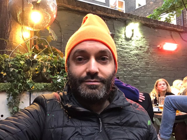 Tired looking bearded man with an orange wooly hat that sticks up a the top like a night cap.