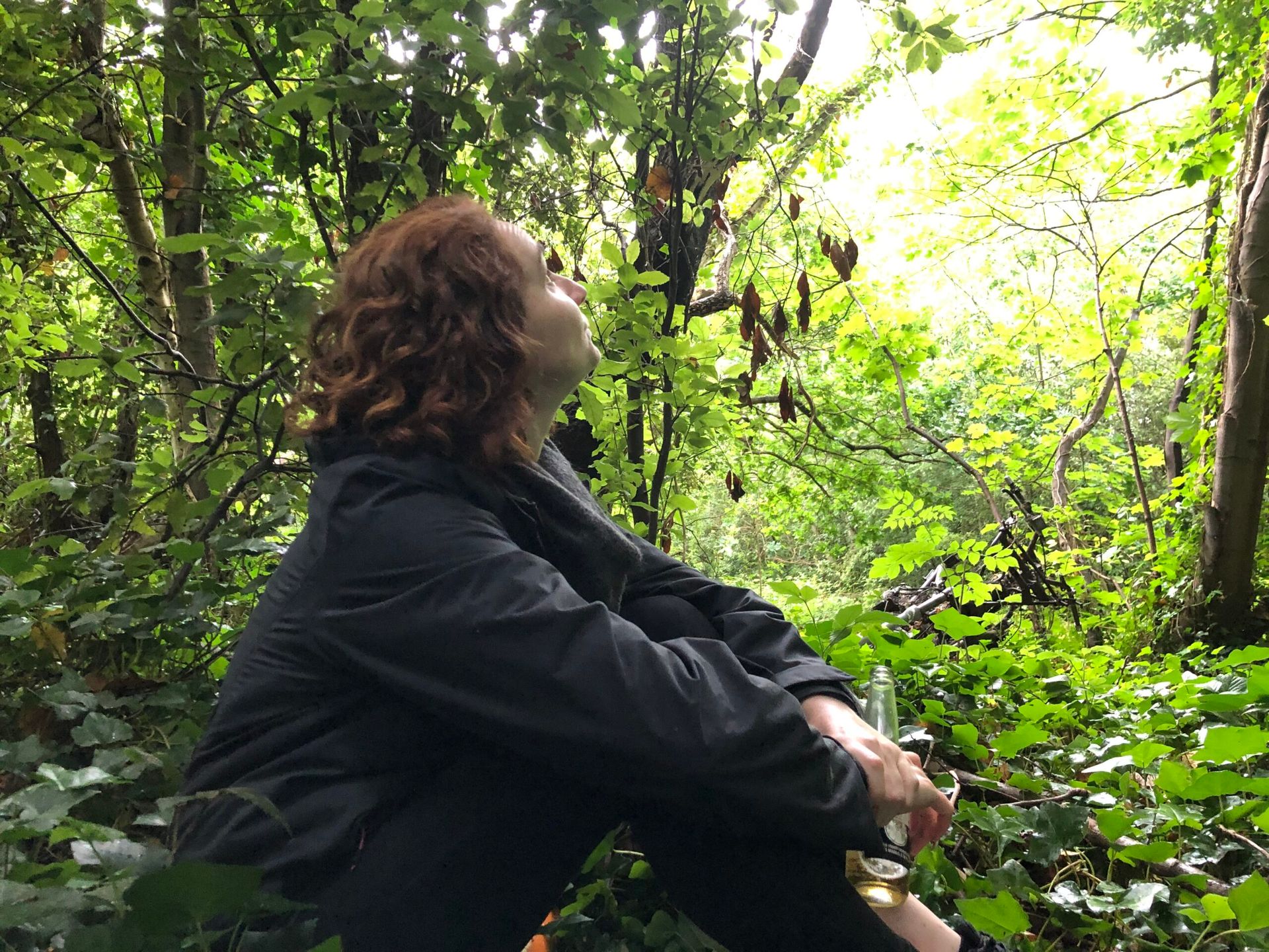 Woman sat in the woods looks up wistfully. She holds a bottle of beer. The remains of a rusted moped can be seen in the background, overgrown with greenery, almost looking like an intentional sculpture