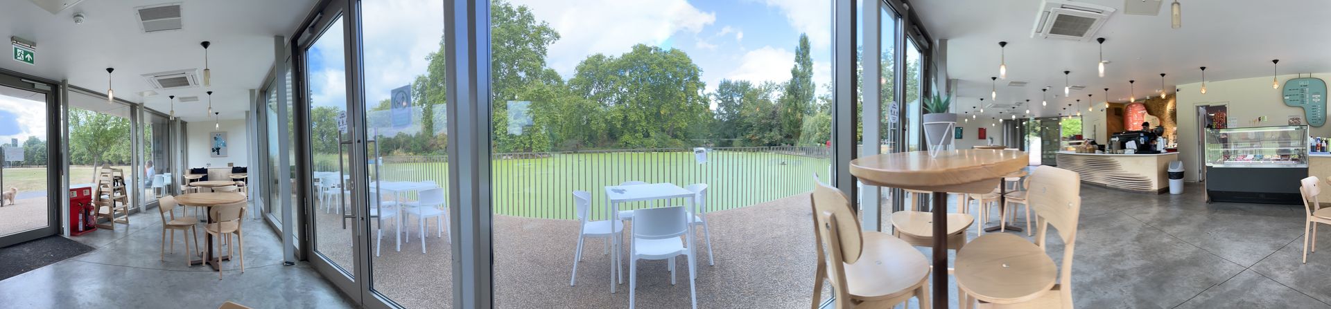 Panorama of a cafe, with glass windows to a lake covered in green algea with trees behind. To the left side is more cafe, with a glass window to a park outside and a small dog. The tables and chairs are wood, bright and clean.